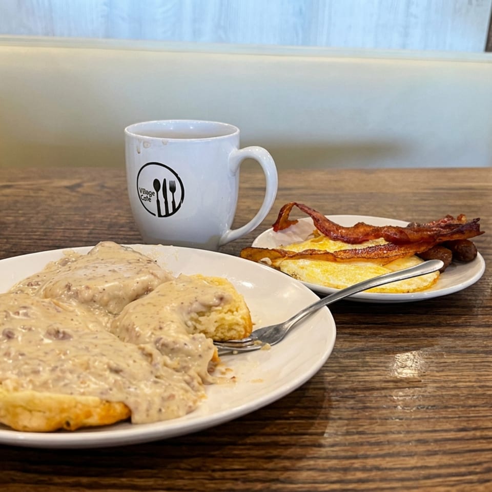 Southern-style breakfast with biscuits and sausage gravy, eggs, bacon, coffee, and a side of creamy grits on a wooden restaurant table.