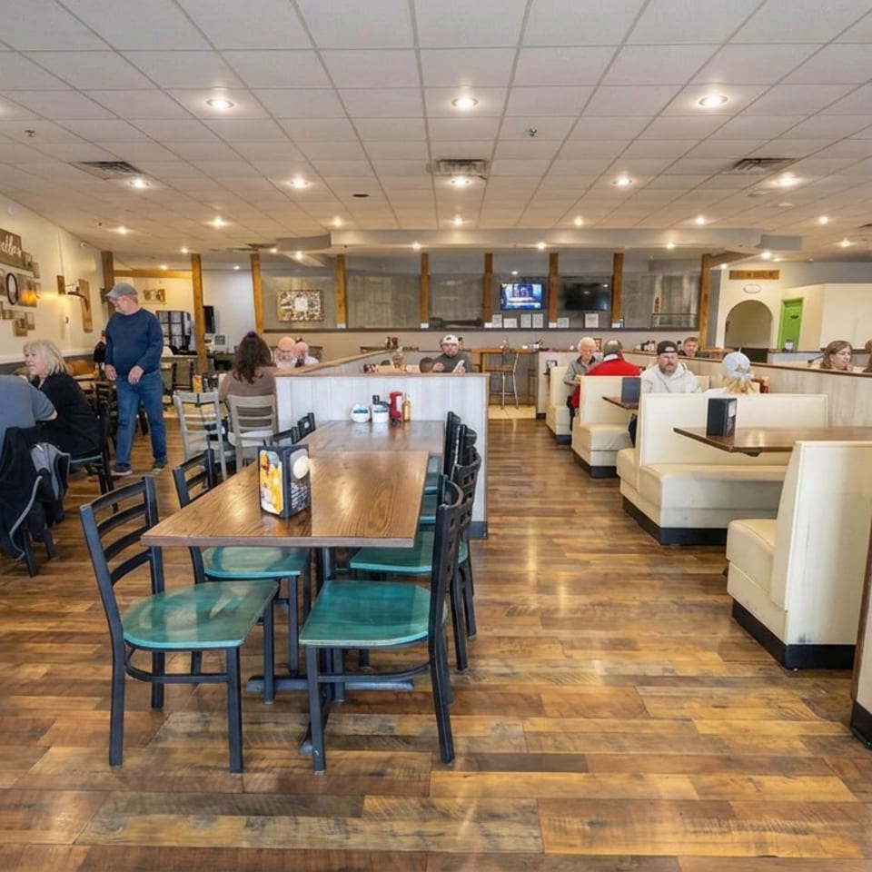 Busy casual restaurant dining room with wooden tables, booths, warm lighting, and patrons dining throughout the space.