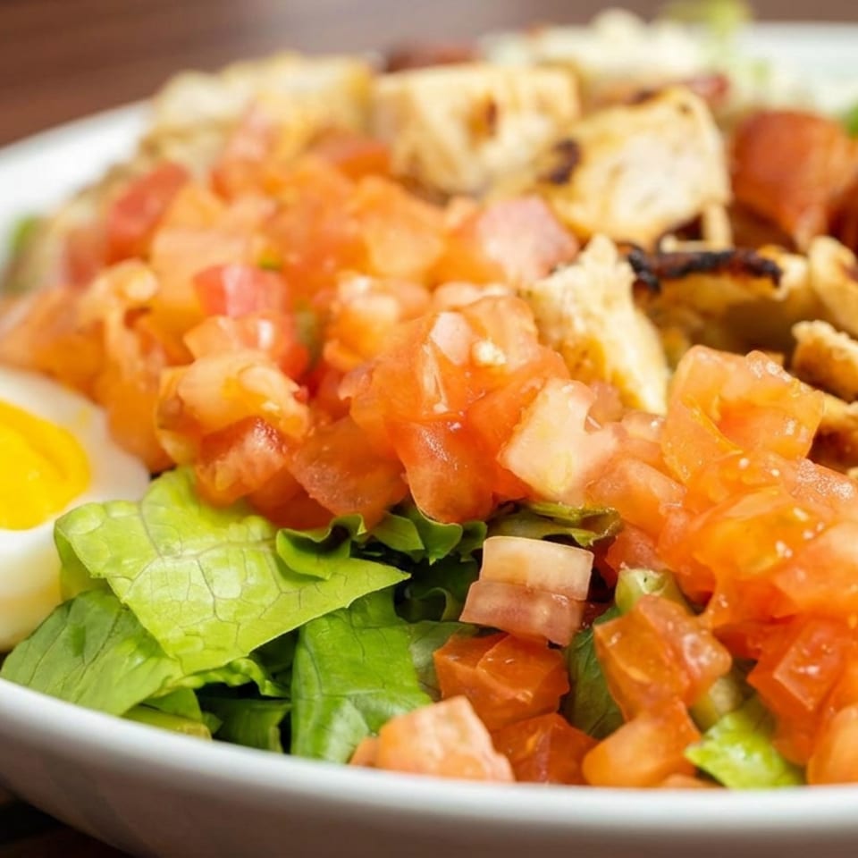 Close-up of a fresh salad with leafy greens, diced tomatoes, grilled chicken, croutons, and a halved boiled egg.