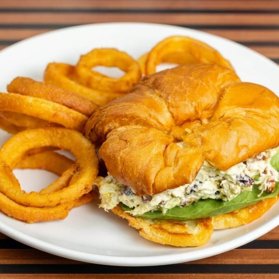 Chicken salad croissant sandwich with lettuce and crispy onion rings on a white plate over a striped wooden table.