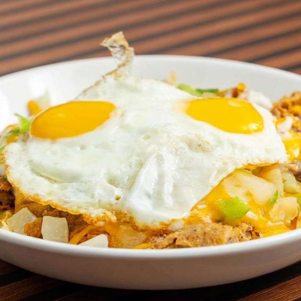 Loaded breakfast bowl topped with two fried eggs, potatoes, vegetables, melted cheese, and seasoned meat on a wooden table.